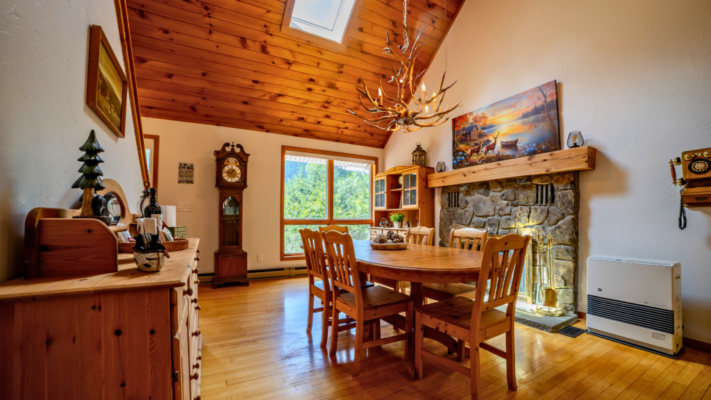 Cozy dining area featuring a wooden table surrounded by chairs, a stone fireplace with a wooden mantle, a large window providing natural light, and rustic decor, including a clock and antler chandelier.