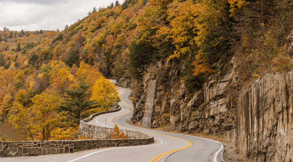 A winding road surrounded by vibrant autumn foliage and rocky cliffs, set against a cloudy sky.