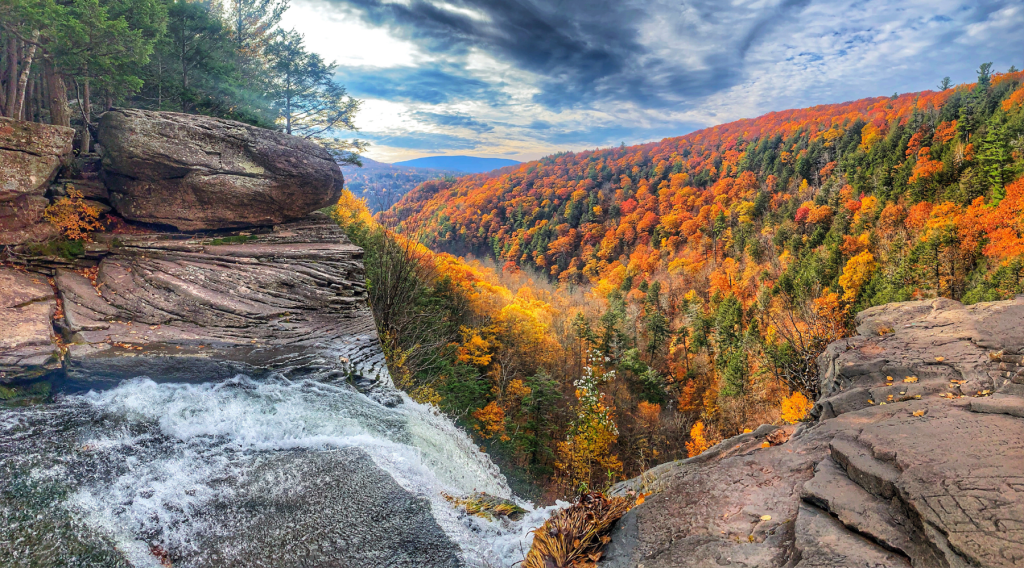 A scenic view from the edge of a rocky cliff overlooking a waterfall, surrounded by vibrant autumn foliage in various shades of orange, yellow, and green, under a partly cloudy sky.
